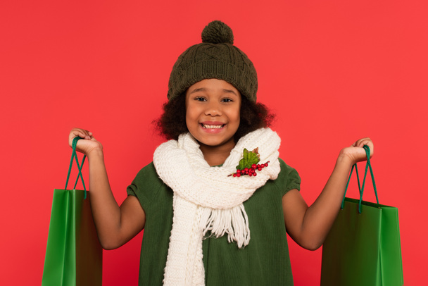 Smiling african american girl in scarf with mistletoe holding shopping bags isolated on red - Foto, Imagem