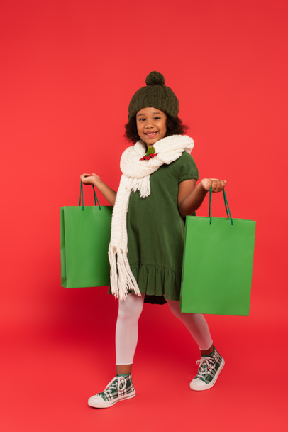 Criança afro-americana com visco no cachecol segurando sacos de compras e andando sobre fundo vermelho - Foto, Imagem