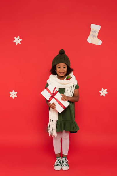 Happy african american girl in knitted hat and scarf with mistletoe holding present near decorative snowflakes on red background - Valokuva, kuva