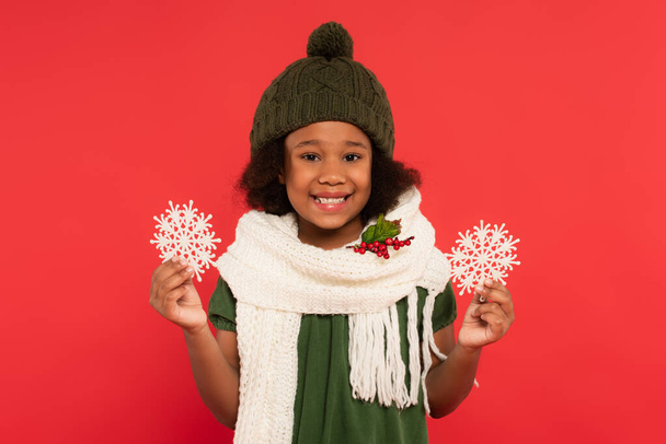 Lächelndes afrikanisch-amerikanisches Mädchen in Mütze und Schal mit Mistelzweigen, die dekorative Schneeflocken isoliert auf rot halten  - Foto, Bild
