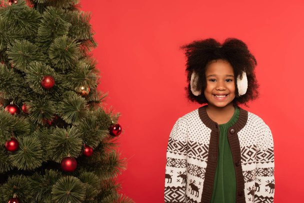 Positive african american kid in ear muffs looking at camera near christmas tree isolated on red  - Photo, Image