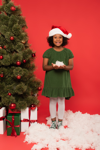 Enfant afro-américain au chapeau de Père Noël tenant de la neige décorative près de l'arbre de Noël et des cadeaux sur fond rouge - Photo, image