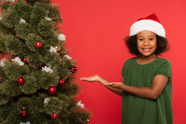 Lächelndes afrikanisch-amerikanisches Kind mit Weihnachtsmütze zeigt auf Weihnachtsbaum auf rotem Grund  - Foto, Bild