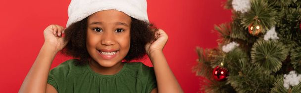 African american kid wearing hat near blurred christmas tree isolated on red, banner  - Photo, Image