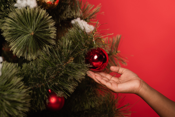 Vista recortada del niño afroamericano sosteniendo la pelota en el árbol de Navidad aislado en rojo  - Foto, imagen