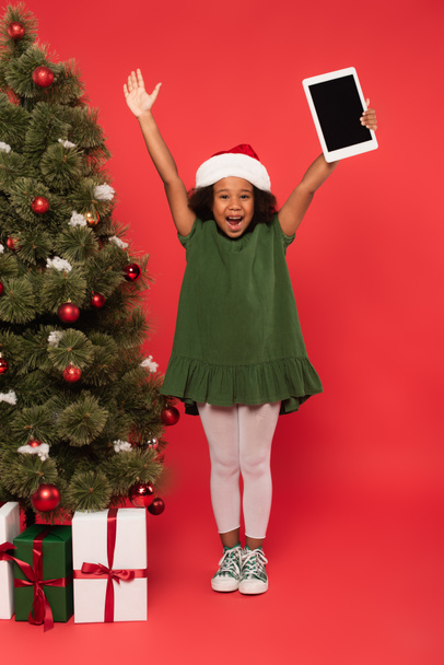 興奮アフリカ系アメリカ人の女の子でサンタ帽子保持デジタルタブレット近くクリスマスツリーとプレゼントオン赤の背景 - 写真・画像