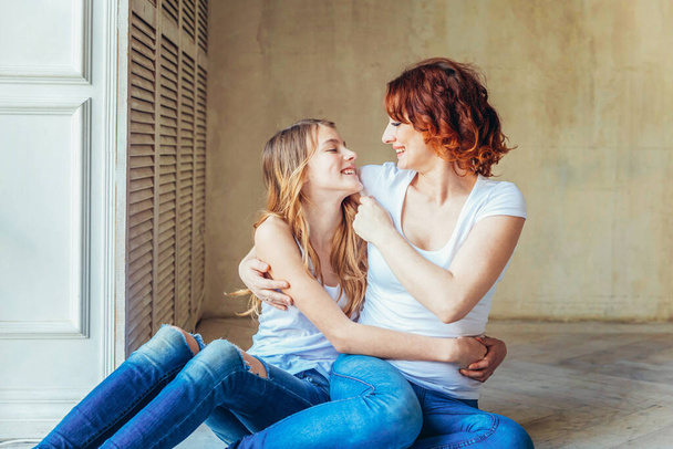 Young mother embracing her child. Woman and teenage girl relaxing in white bedroom near gray wall indoors. Happy family at home. Young mom playing with her daughter - Foto, Imagem
