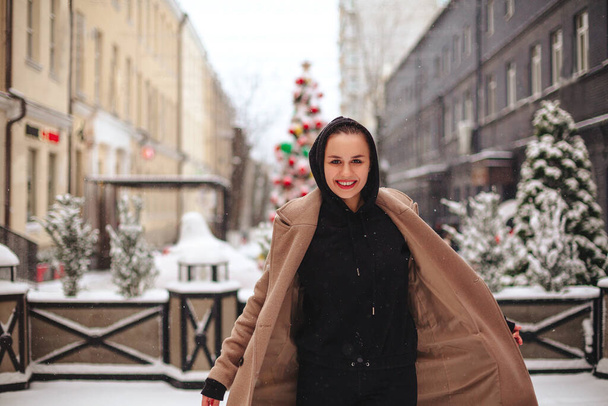 Giovane donna positiva con un sorriso divertente sul suo viso si trova all'esterno in cappotto beige e sciarpa nera sulla testa, tremando dal freddo invernale sullo sfondo di alberi ricoperti da uno spesso strato di neve. - Foto, immagini