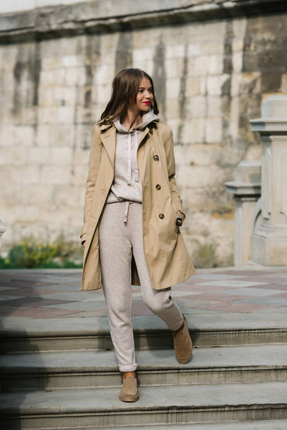 Portrait of fashionable women in beige sports suit, trench coat and stylish suede loafer posing on the stone stairs - Foto, Imagen