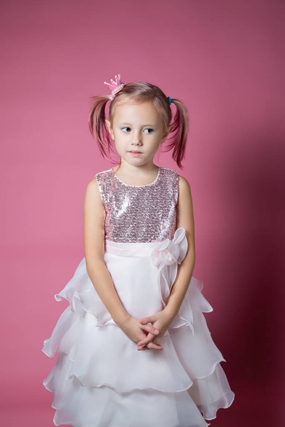 Little caucasian girl in a festive dress with sequins posing like a princess on a pink background looking at the camera - Photo, image