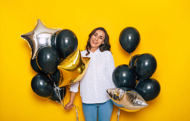 Smiling Happy Excited cute girl holding many black balloons in hands is posing on the yellow background while shopping on Black Friday - 写真・画像