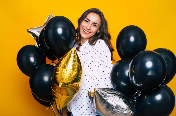 Smiling Happy Excited cute girl holding many black balloons in hands is posing on the yellow background while shopping on Black Friday - Foto, imagen