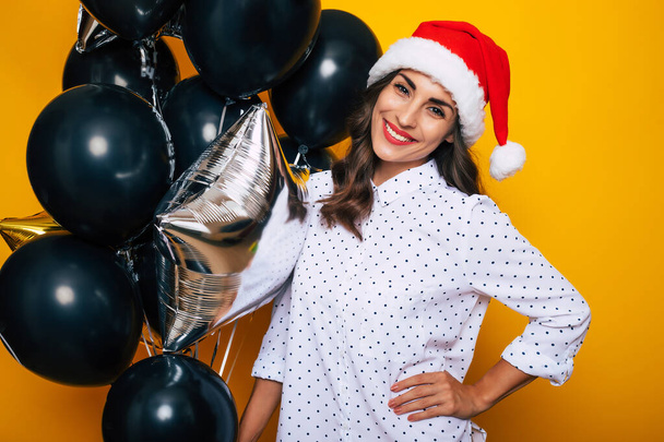 Cute excited young woman in a red Santa hat with air helium balloons posing on the isolated yellow background while celebrating a Christmas and New Year - 写真・画像