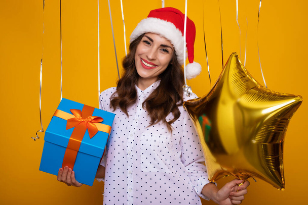 Smiling beautiful woman in a red Santa hat with a gift box in hand is posing on the isolated yellow background while celebrating of Christmas and New Year - Valokuva, kuva