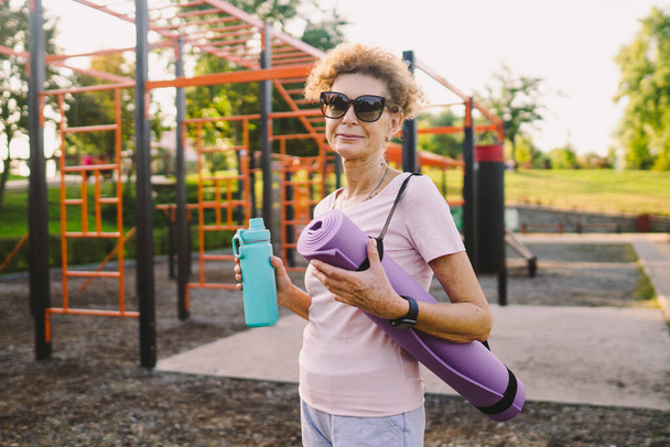 Mature woman going to yoga class outdoors. Active senior enjoying healthy lifestyle. Senior female holding exercise mat while walking at training session. Positive elderly woman with sports equipment. - Photo, Image