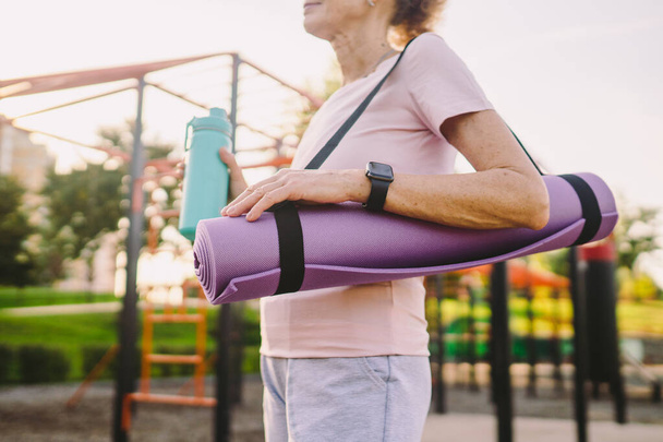 Aktív idős nő fitneszszőnyeggel és vizes palackkal a kezében, ahogy az edzőteremben sétál. Boldog érett hölgy sétál a sportpályán megy a szabadban jóga órán. Aktív életmód koncepció. - Fotó, kép