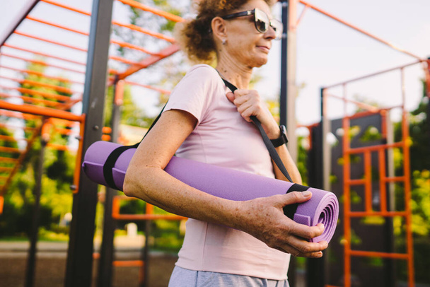 Felnőtt nő, aki jóga órára megy a szabadban. Aktív nyugdíjas élvezi az egészséges életmódot. Végzős női lábtörlő lábtörlővel séta közben az edzésen. Pozitív idős nő sportfelszereléssel. - Fotó, kép