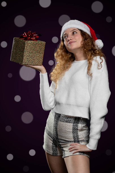young latin woman showing a christmas present with a santa claus hat. space for text - Фото, изображение