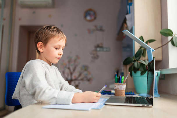 Cute little schoolboy studying at home doing school homework. Training books and notebook on the table. Distance learning online education - 写真・画像