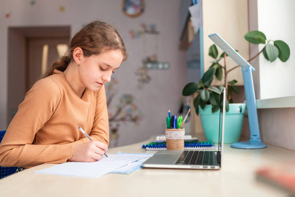 Beautiful schoolgirl studying at home doing school homework. Training books and notebook on the table. Distance learning online education - Photo, Image