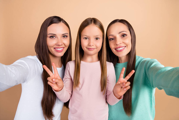Self-portrait of three beautiful trendy cheerful girls showing v-sign isolated over beige pastel color background - 写真・画像