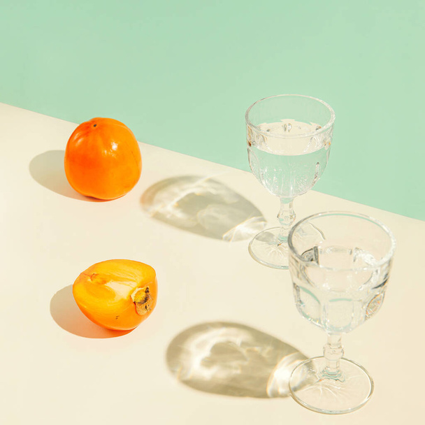 Persimmon ovoce a sklenice vody na geometrickém pastelovém pozadí se sluncem. Letní nápoje a koncept občerstvení. Minimální složení stylu - Fotografie, Obrázek