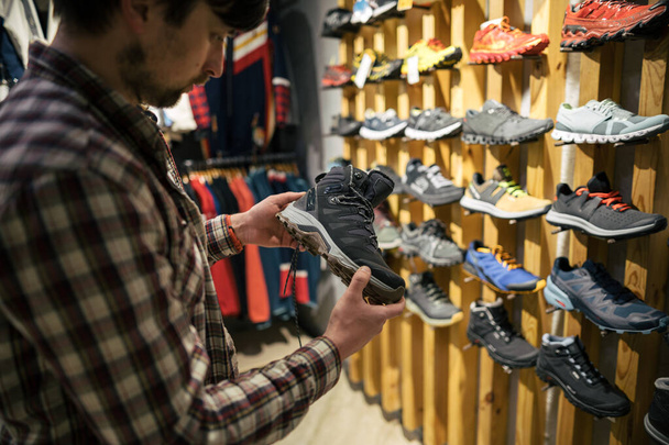 Kaukaski mężczyzna trzyma i bada buty turystyczne przed zakupem w sklepie dla turystyki i sportu. Klient ocenia jakość obuwia turystycznego w sklepie. Wodoodporne wędrówki zimą jesienne buty. - Zdjęcie, obraz