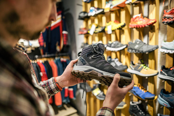 Caucasian man chooses and measures hiking boots at a sports and travel store. Man looking at hiking shoes in outdoor shop. Male shopping hiking boots in sports shop. Travel goods store theme. - Photo, Image