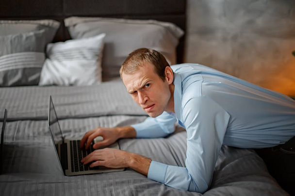Похмурий чоловік підняв брову і з обуренням виглядав. позаштатний бізнесмен працює на ноутбуці, що сидить біля ліжка в будинку. Збентежений бізнесмен
 - Фото, зображення