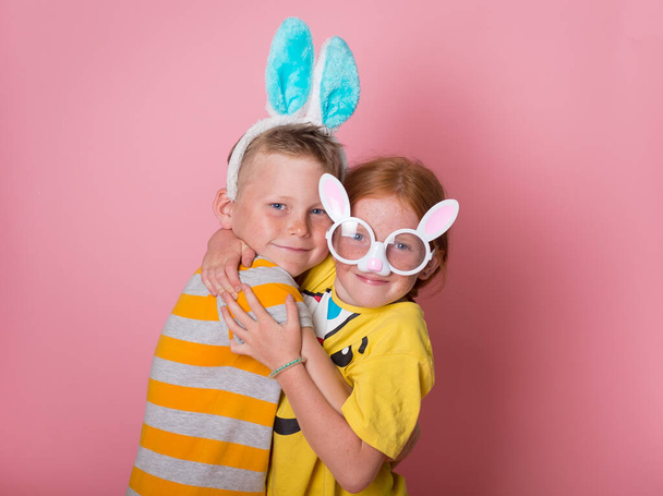 Веселі щасливі діти на полюванні на великодні яйця. Шкільні діти з вухами кролика обіймаються один з одним. Блондинка хлопчик і руде волосся дівчина в костюмі кролика грають на рожевому фоні
. - Фото, зображення