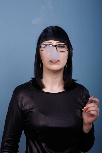 Een mooi meisje met donker haar en bril rookt sensueel een elektronische sigaret op een blauwe achtergrond - Foto, afbeelding