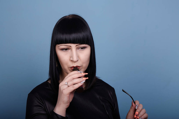 Ένα όμορφο κορίτσι με σκούρα μαλλιά και γυαλιά καπνίζει αισθησιακά ένα ηλεκτρονικό τσιγάρο σε μπλε φόντο - Φωτογραφία, εικόνα
