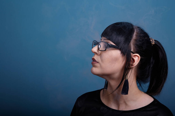 A beautiful girl with dark hair and glasses exquisitely smokes an electronic cigarette on a dark background - Fotografie, Obrázek