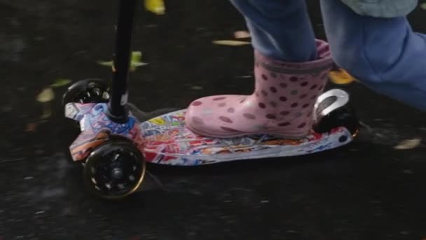 Dziewczynka w gumowych butach jeździ po mokrym asfalcie pokrytym kałużami i spadającymi liśćmi na skuterze. Full HD slow motion zbliżenie wideo. - Materiał filmowy, wideo