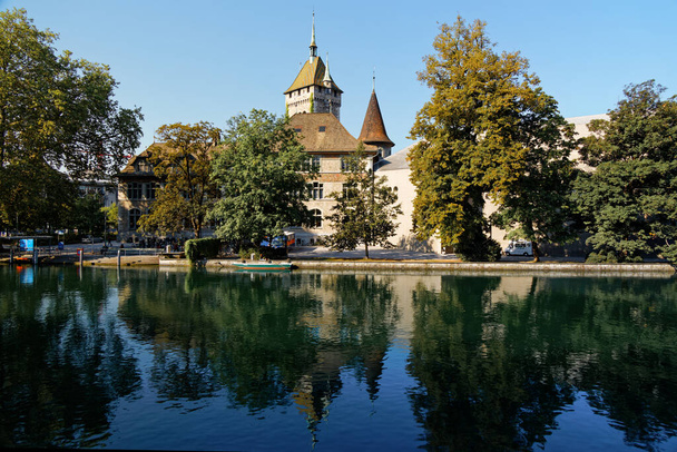 国立風景博物館秋の朝、チューリッヒ、青い空の前の木々の間の建物や尖塔、クローズアップビュー、川の前景 - 写真・画像
