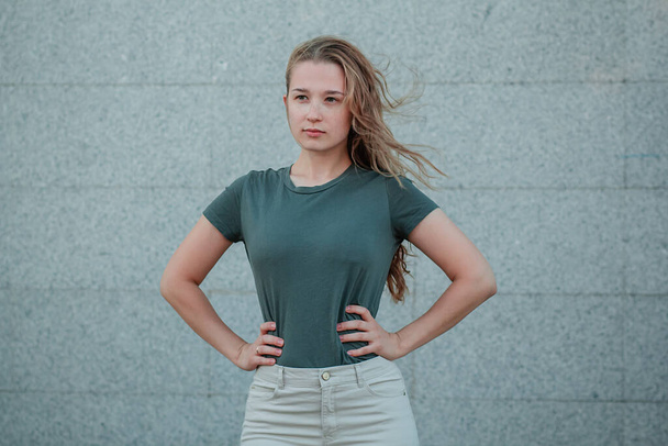 A young woman in a dark turquoise T-shirt stands against a gray wall in the street. Girl in empty clothes. Mock-up. - Foto, immagini