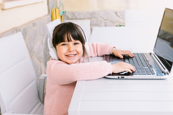 Kleines Mädchen mit weißen Kopfhörern, rosa Pullover, vor dem Laptop sitzend, tippend, auf der Terrasse, lächelnd. Konzept des Fernstudiums Online, Kindheit und Technik. - Foto, Bild