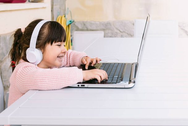 Маленькая девочка в белых наушниках, в розовом свитере, сидит перед ноутбуком, печатает и двигает мышкой, на террасе, улыбается. Дистанционное образование, детство и технологии онлайн. - Фото, изображение