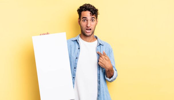 hispanic handsome man looking shocked and surprised with mouth wide open, pointing to self. empty canvas concept - Foto, Bild