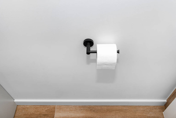 Suporte preto fosco com papel higiênico preso à parede do vaso sanitário, um piso coberto com telhas cerâmicas imitando madeira. - Foto, Imagem