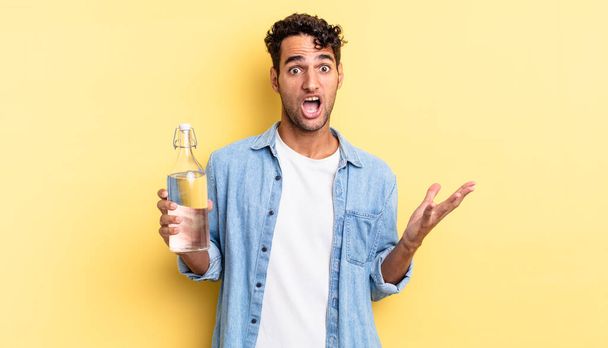 hispanic handsome man amazed, shocked and astonished with an unbelievable surprise. water bottle concept - Zdjęcie, obraz