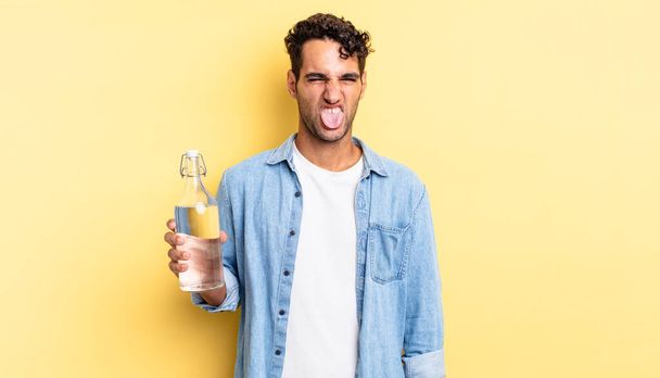Латиноамериканец, красивый мужчина, чувствует отвращение, раздражение и высунутый язык. бутылка с водой - Фото, изображение