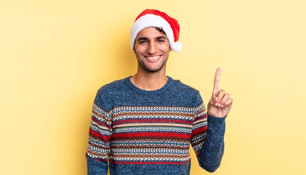 hispanic handsome man smiling and looking friendly, showing number one. christmas concept - Foto, Bild