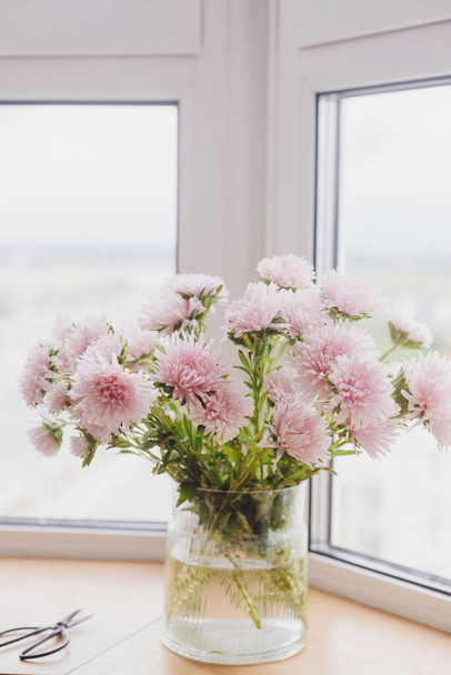 Beautiful autumn flowers and scissors in light on wooden windowsill. Pink asters flowers at big windows in modern room. Floral decoration for fall holidays in countryside home. Cozy autumn - Photo, Image