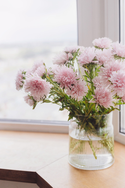 Beautiful autumn flowers in light on wooden windowsill. Pink asters flowers at big windows in modern room. Floral decoration for autumn holidays. Cozy home in fall season - Photo, Image