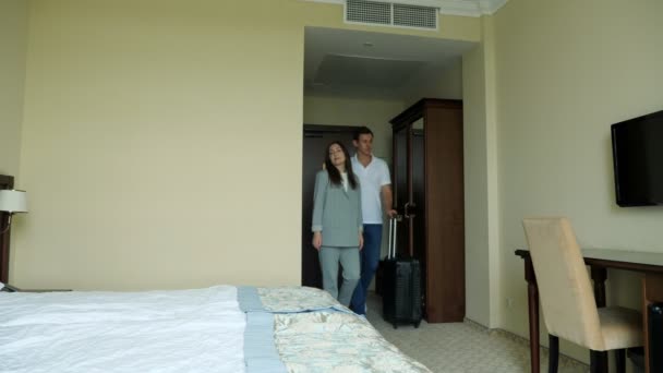 mladý pár s kufrem vejdou do hotelového pokoje a posadí se na postel - Záběry, video