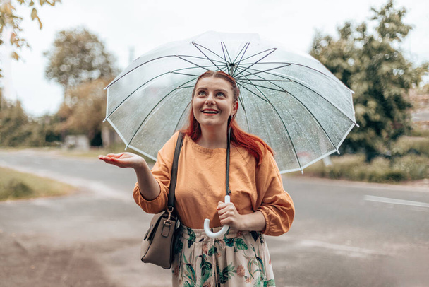 Krásná dívka stojí pod průhledným deštníkem, natahuje ruku a chytá kapky vody. Vodorovný nápis, kopírovací prostor - Fotografie, Obrázek