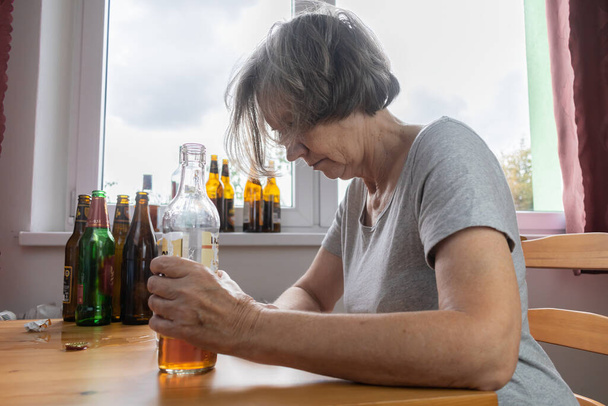 стара старша жінка сидить поруч зі столом п'є пляшку алкоголю вдома сумно самотній алкоголізм Ознаки та симптоми зловживання та проблеми зі одужанням
 - Фото, зображення