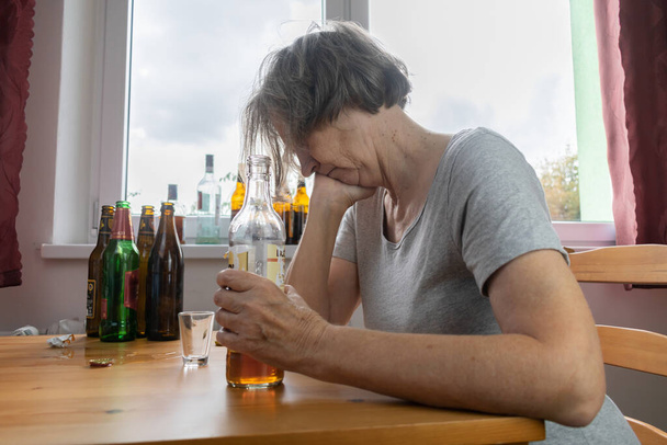 Yaşlı bir kadın masanın yanına oturur evde yalnız başına alkol şişesi içer üzgün alkolizm belirtileri ve iyileşme sorunları yaşar. - Fotoğraf, Görsel