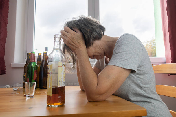 alte Seniorin sitzt am Tisch und trinkt zu Hause eine Alkoholflasche, traurig allein Alkoholismus Anzeichen und Symptome Reha-Missbrauch und Genesungsprobleme - Foto, Bild